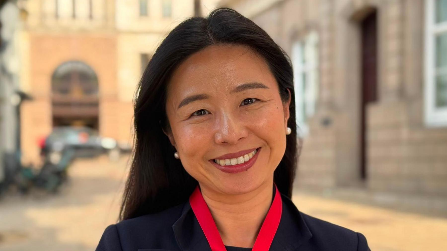 Victoria Li in a dark blazer standing on a town street, with historic buildings blurred in the background.