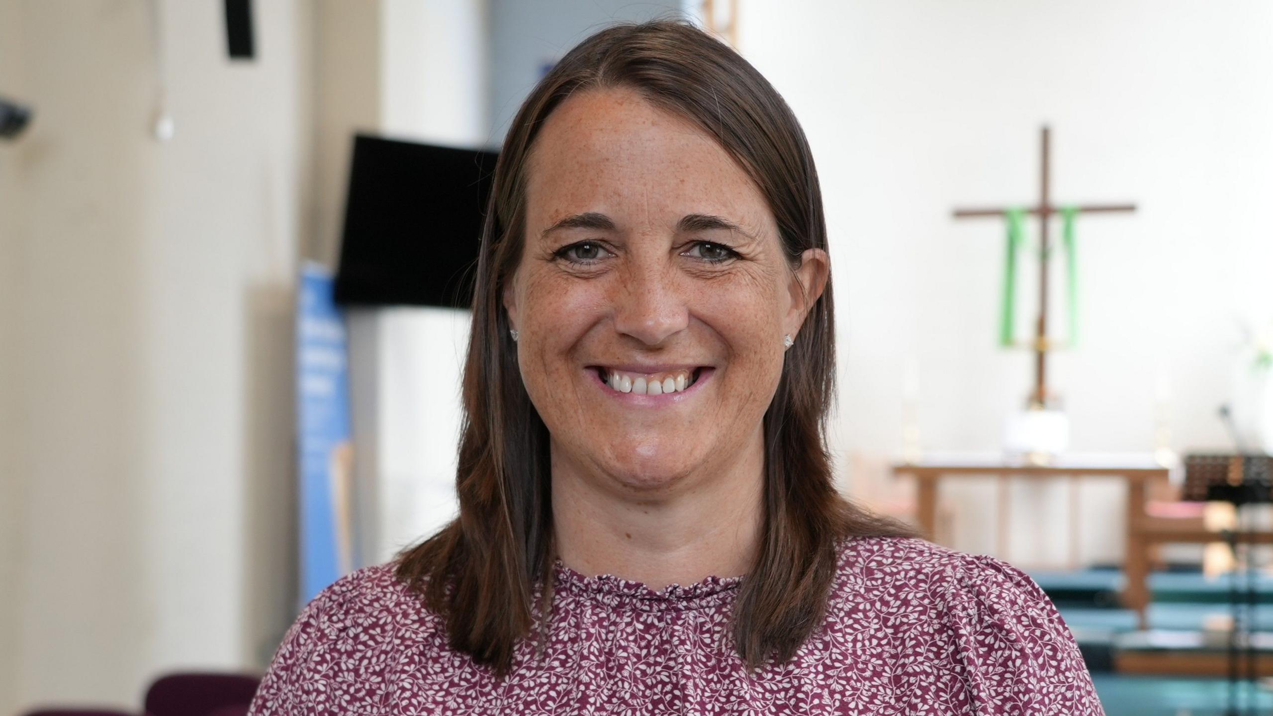 A woman with shoulder-length hair smiles at the camera. She has brown hair and brown eyes and is wearing a burgundy and white patterned top. Behind her is a simple altar with a wooden table and a cross displayed on a white wall.