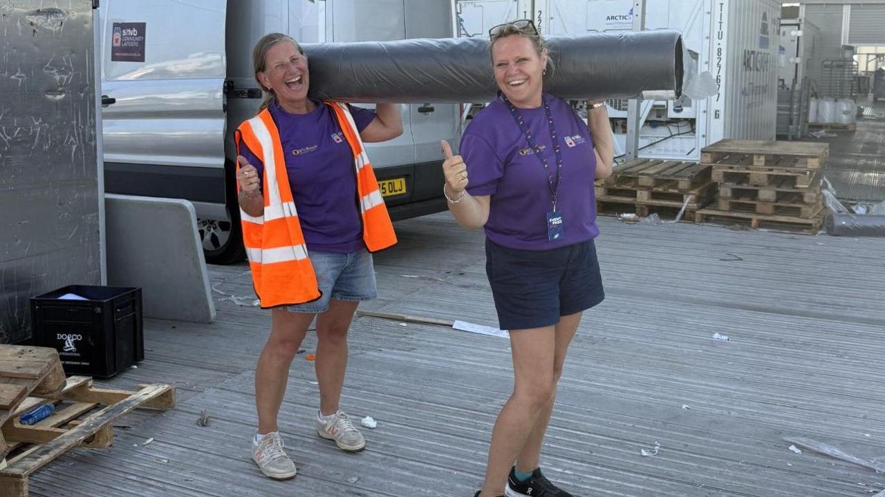 Two women, both holding their thumbs up, holding a carpet roll, both wearing shorts, and a purple T-shirt. The one on the left has on a high-vis jacket. A silver van is behind them and some wooden pallets.