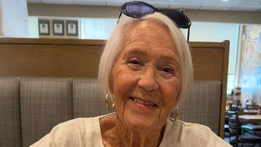 Gloria Stephenson, seated in a restaurant and smiling at the camera. She has short, white hair and is wearing pink lipstick, gold hoop earrings and a white top.