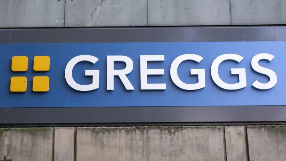 A Greggs bakery sign has white lettering spelling out the company name with an orange logo of four squares on the blue background.