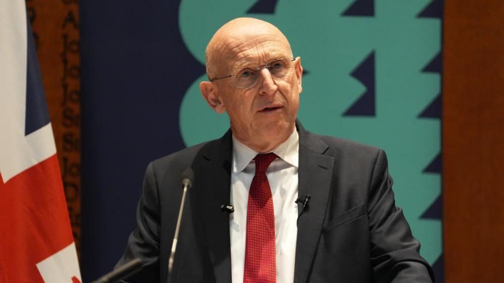 John Healey wearing a dark suit and red tie standing before a microphone