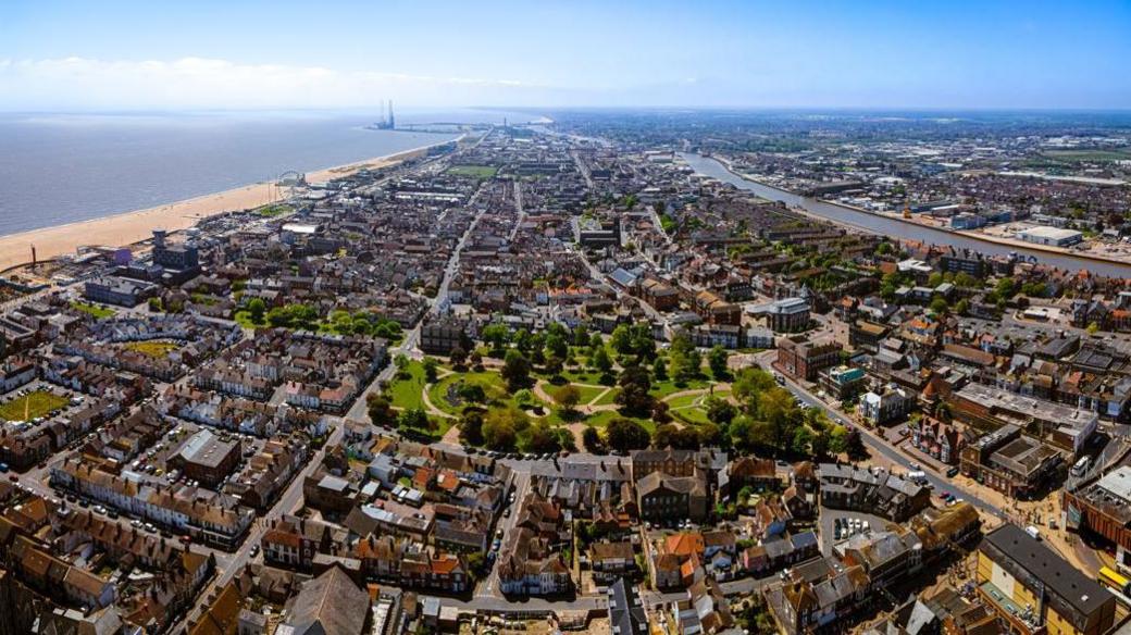 A bird's eye view of Great Yarmouth which includes areas of dense housing and a park, with the beach and the sea to the left. It is a sunny day and the sky is blue.