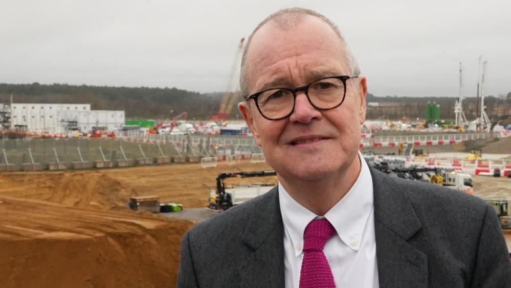 Sir Patrick Vallance is standing in front of a large construction site, with cranes and piles of dirt in the background. He is wearing a grey suit, white shirt and dark red tie.