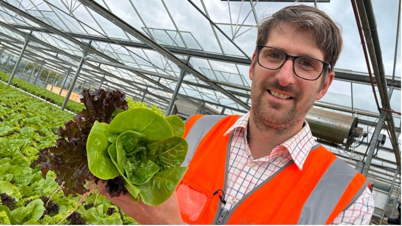 Mathew Brankley stands to the right of the image wearing a checked shirt and a high-vis waistcoat. He is holding a lettuce. Behind him is a crop of salad leaves.