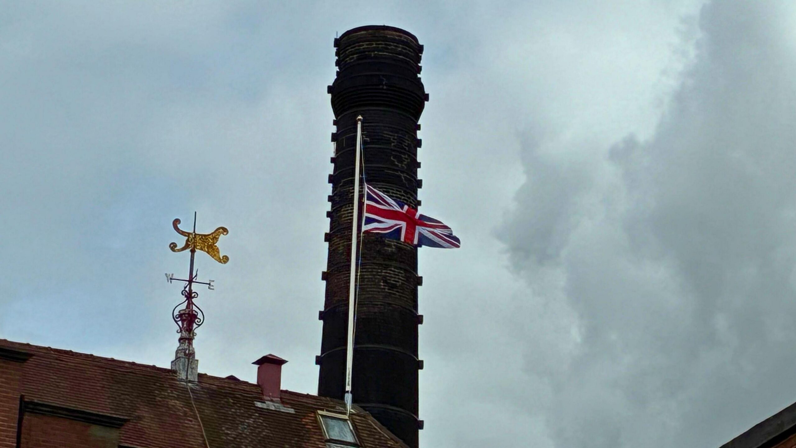 A Union Jack flag on top of a brewery flies at half mast against a cloudy grey sky.