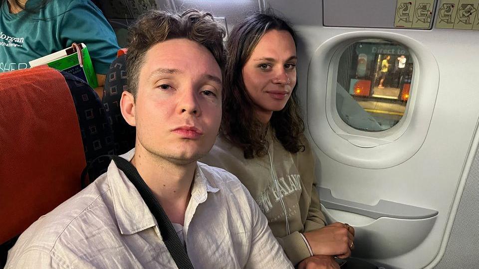 Sammy and Raiff on the diverted EasyJet plane. They are both looking in to the camera. Faiff wears a white shirt and Sammy wears a light green jumper.