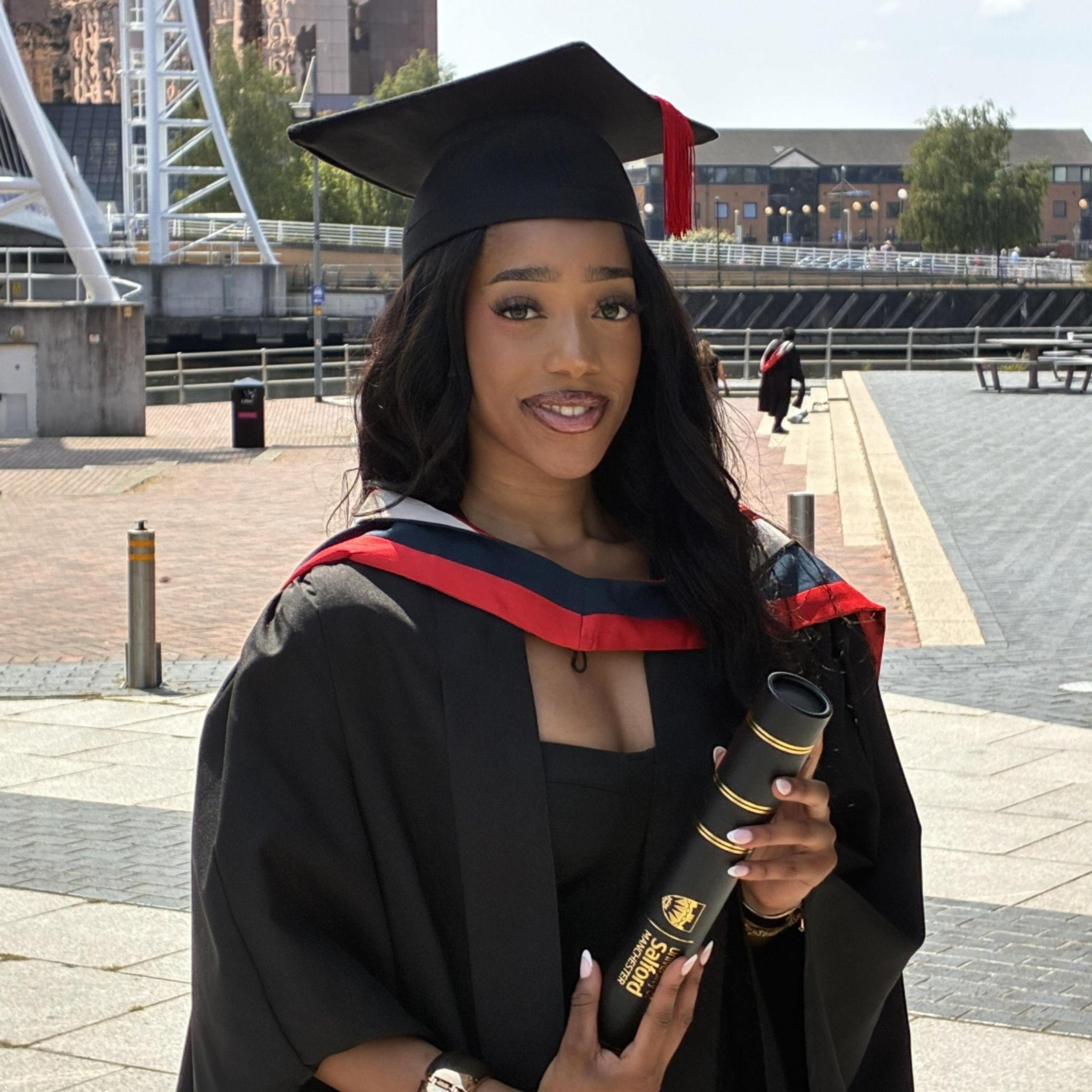 A woman wearing a black dress with a black gown and mortar board over the top, stands as she holds her degree. It is rolled and in a tube emblazoned with the name of her university and its logo. A bridge over a river and a tall office building can be seen in the background.
