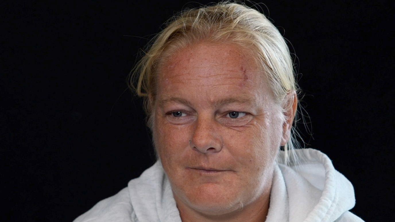 A woman with blonde hair scraped back looks off camera. She is wearing a white hoodie. The background is black.