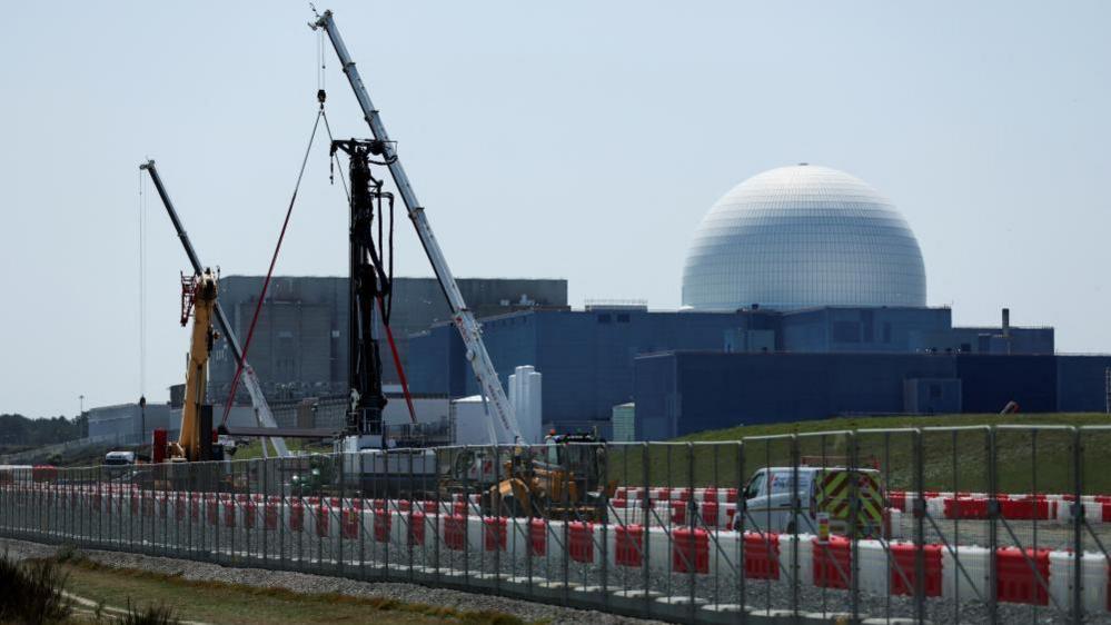 The large white dome of the Sizewell B nuclear power plant is the backdrop of the picture which has construction work and cranes in the foreground. The white dome sits on large square grey coloured buildings.