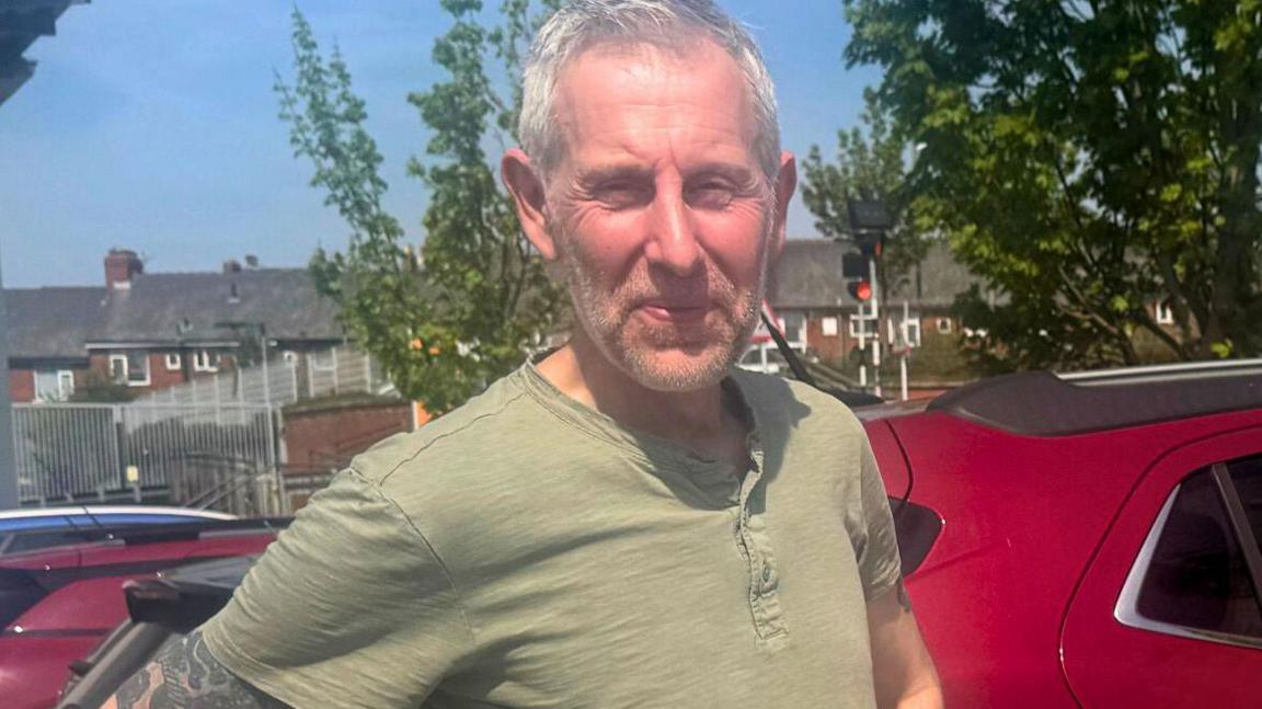 A man with short grey hair and a khaki t-shirt stands in a car park.