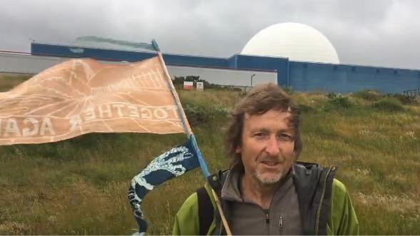 A man stands in front of a large power station on a grass bank. He has medium length brown hair and is wearing a green coat and brown jumper. He is holding an orange flag.