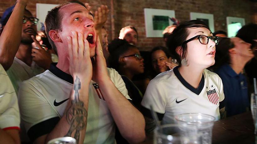 Two United States football fans react during the match between Ghana and USA at the WoodWork bar in the Brooklyn borough of New York City. One is a man and the other is a woman. They are both wearing USA football tops.