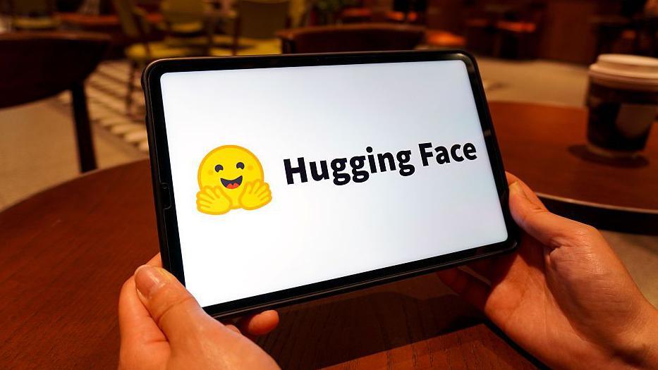The Hugging Face logo shown on a tablet screen. It is a yellow emoji of a smiling face with its hands spread out in front of it.