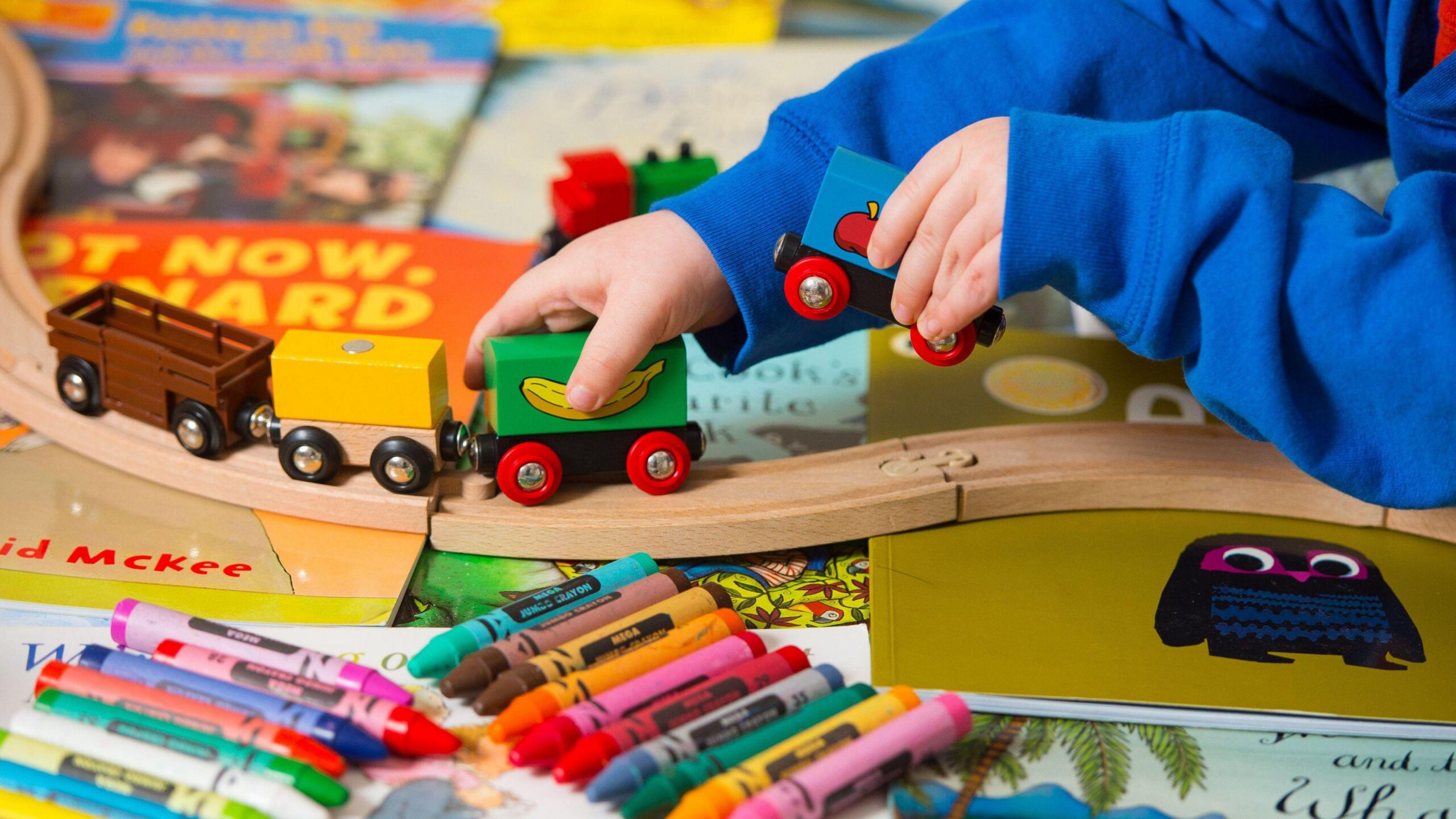 A toddler playing with a toy train set which is sat on top of colourful books and crayons in front of the train line.