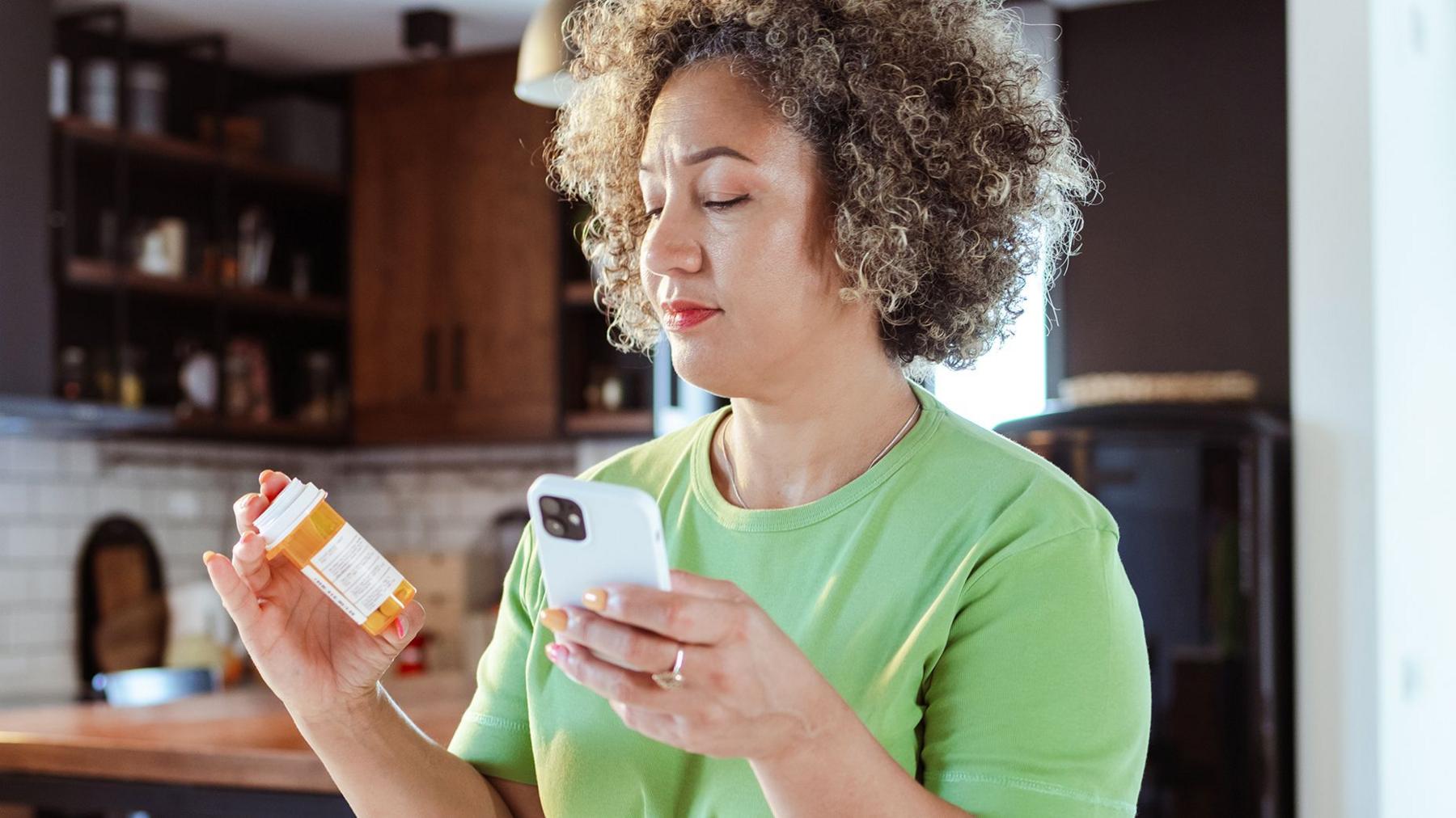 A woman wearing a green t-shirt stands in her kitchen holding a bottle of pills in one hand and her phone in the other - she is looking at the bottle of pills