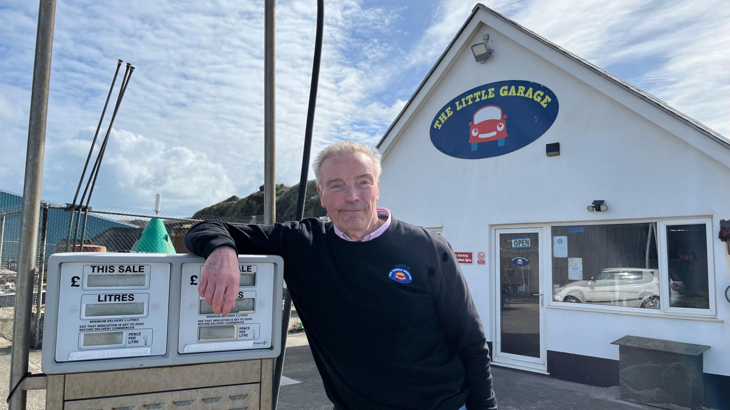 Dave Cranwell, leaning on a petrol pump infront of his his garage, which has a logofor "The Little Garage" which depicts a smiling car. Dave has short grey hair, and is wearing a jumper which also has a small logo on it.