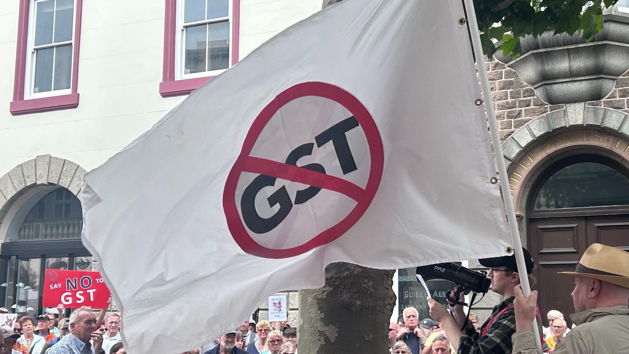 A crowd of people gather in a town square. In the foreground is a white flag containing a red circle with a red line through it, like a traffic sign. The letters "GST" are scored through by the line