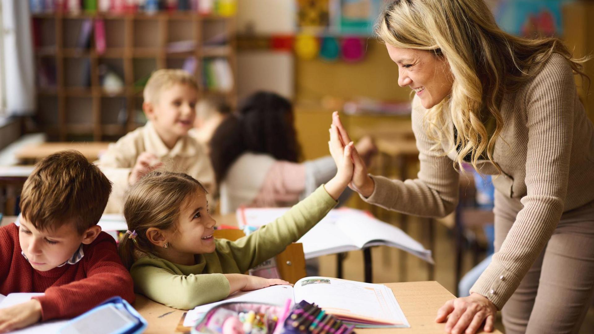 A young teacher is leaning over the desk of two under-10 pupils and is high-fiving the nearest child. Both are smiling.