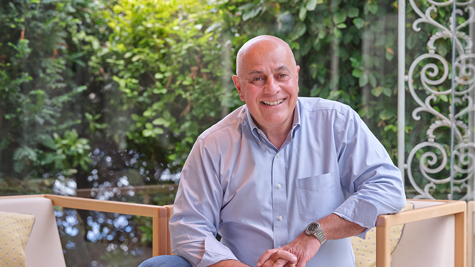 A bald man in a blue shirt sits on a cream chair on a veranda, with a hedge and cream ironwork pillar visible behind him. He is smiling.
