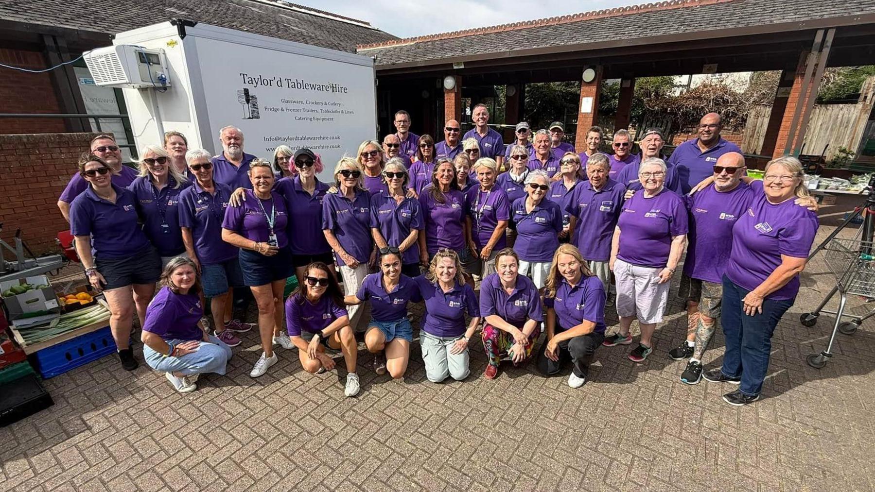 A large group of volunteers, all wearing purple T-shirts with six kneeling at the front and the rest standing behind them. They are standing next to a white van and there is a brick building behind them. A shopping trolley is to the right.