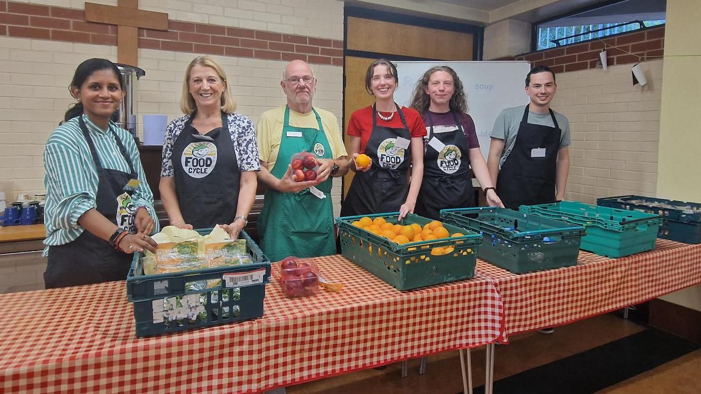 FoodCycle volunteers pose behind tables with crates of oranges, apples, and other produce while wearing aprons in a community hall, ready to prepare food for a community meal. Ian White is stood with them with glasses, a yellow t-shirt and green apron.