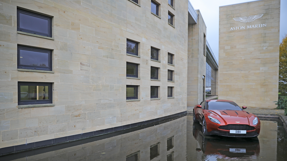 A red Aston Martin car is displayed outside the company's manufacturing site in Warwickshire. The building has white bricks and Aston Martin plus its logo displayed on it in silver.