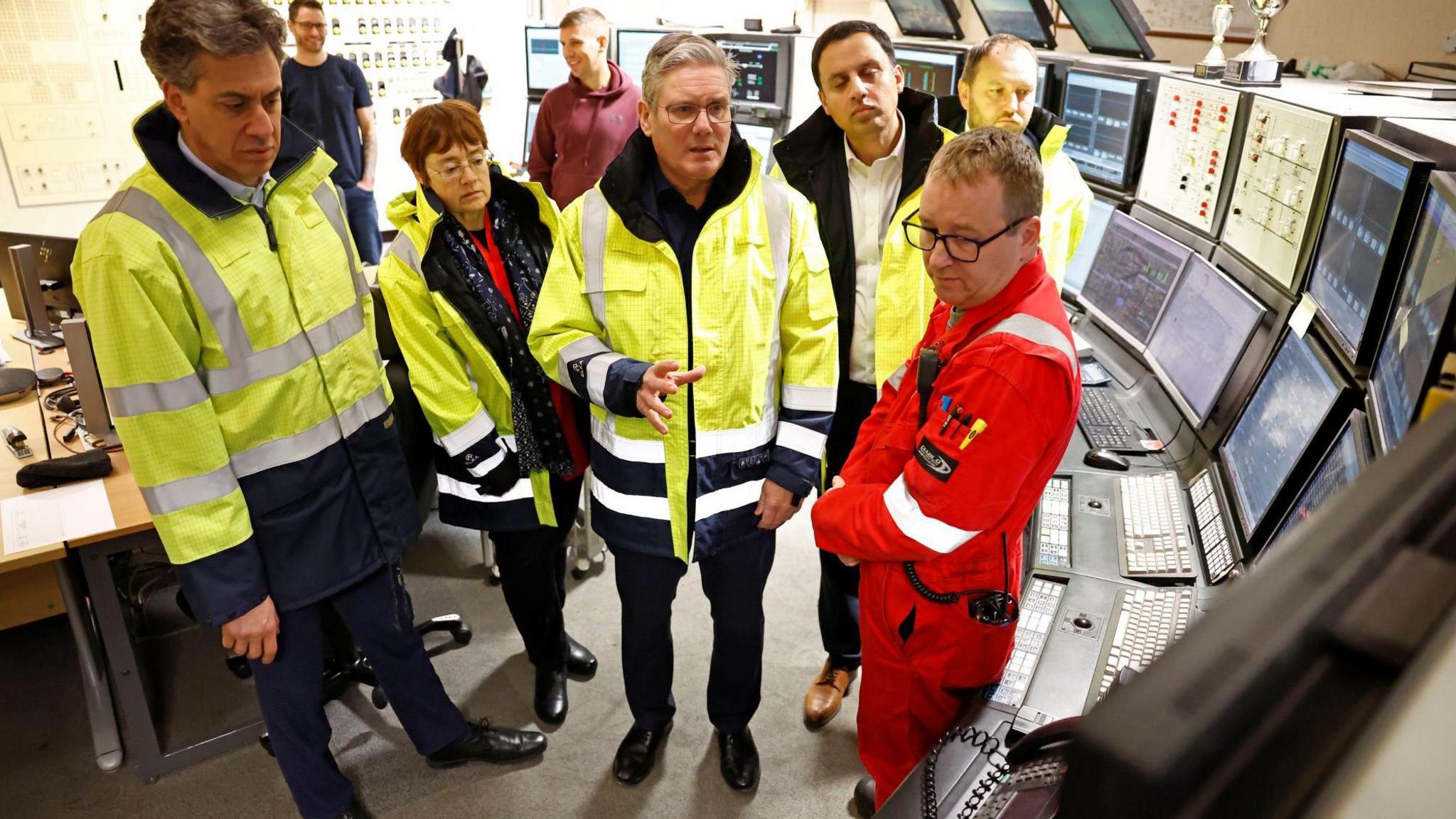 Ed Miliband, Sarah Boyack, Sir Keir Starmer, Anas Sarwar and Ian Murray wear an array of hi-vis jackets while staring at a bank of control screens. A man is speaking to them. They are in a control room.