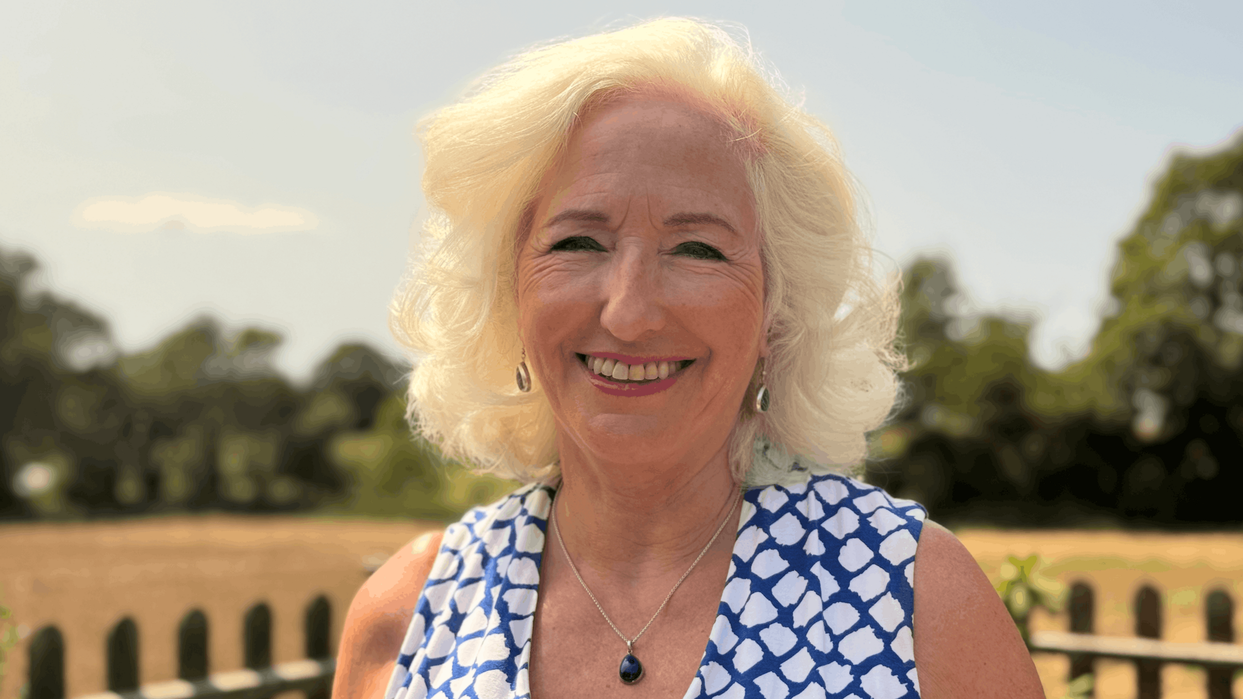 Debbie Taylor wearing a blue-and-white patterned sleeveless top, standing outdoors in front of a fence, field and trees.