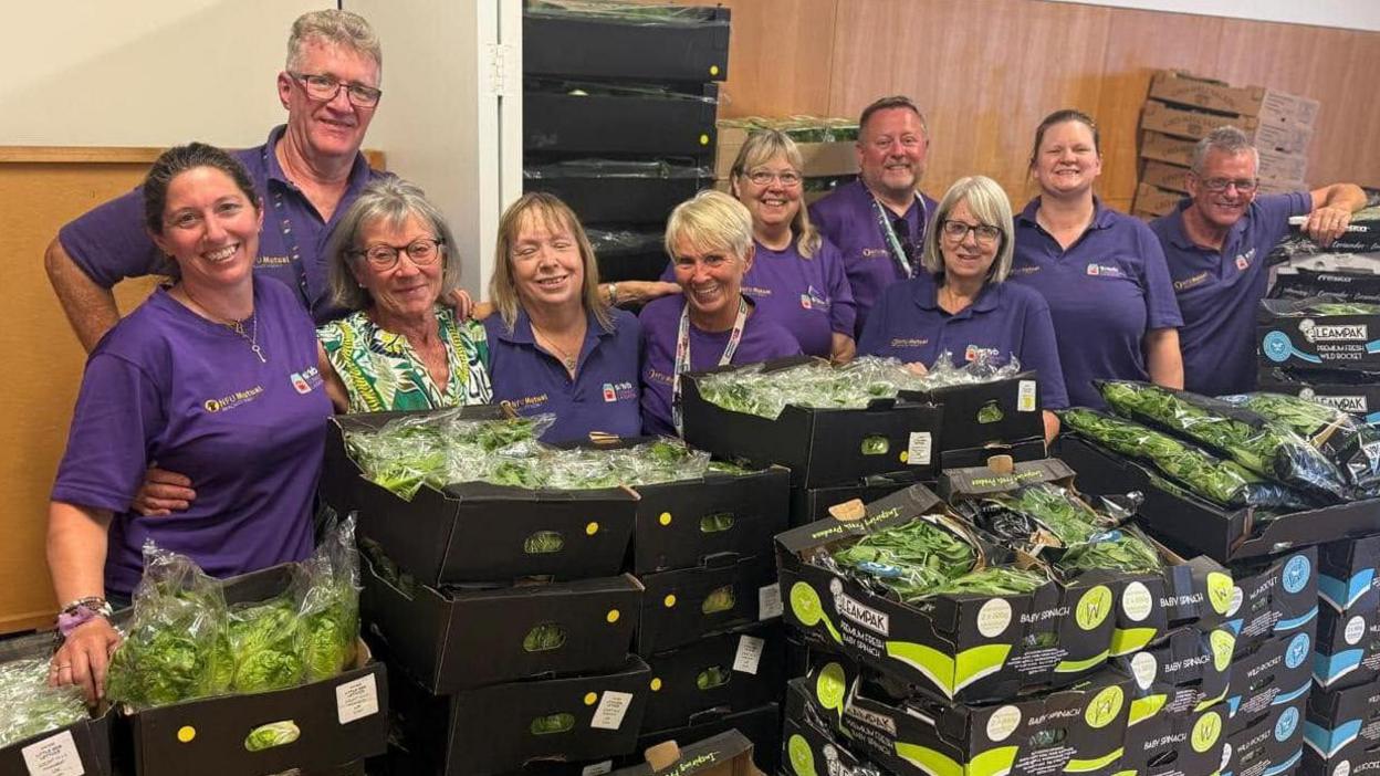 A large group of people, in standing behind a large selection of boxes of salad. They are all wearing purple T-shirts. They are in a room with items all behind them and are all smiling and looking at the camera.
