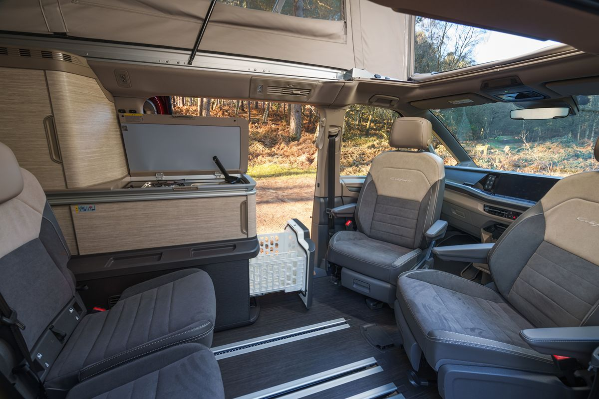 Interior of a modern camper van with swivel front seats, pop-up roof, and a compact kitchen unit with an open lid.