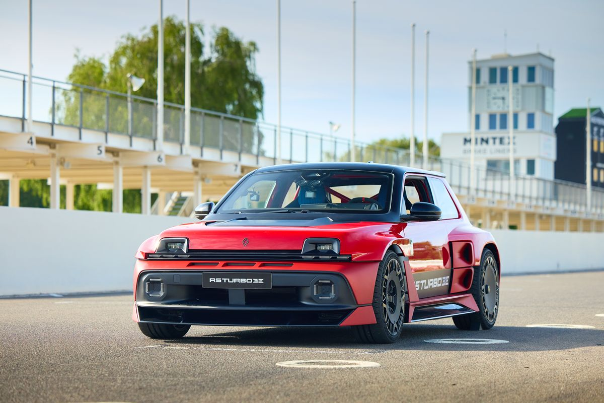 Red and black Renault R5 Turbo 3E concept car on a racetrack, with grandstands and a control tower in background.